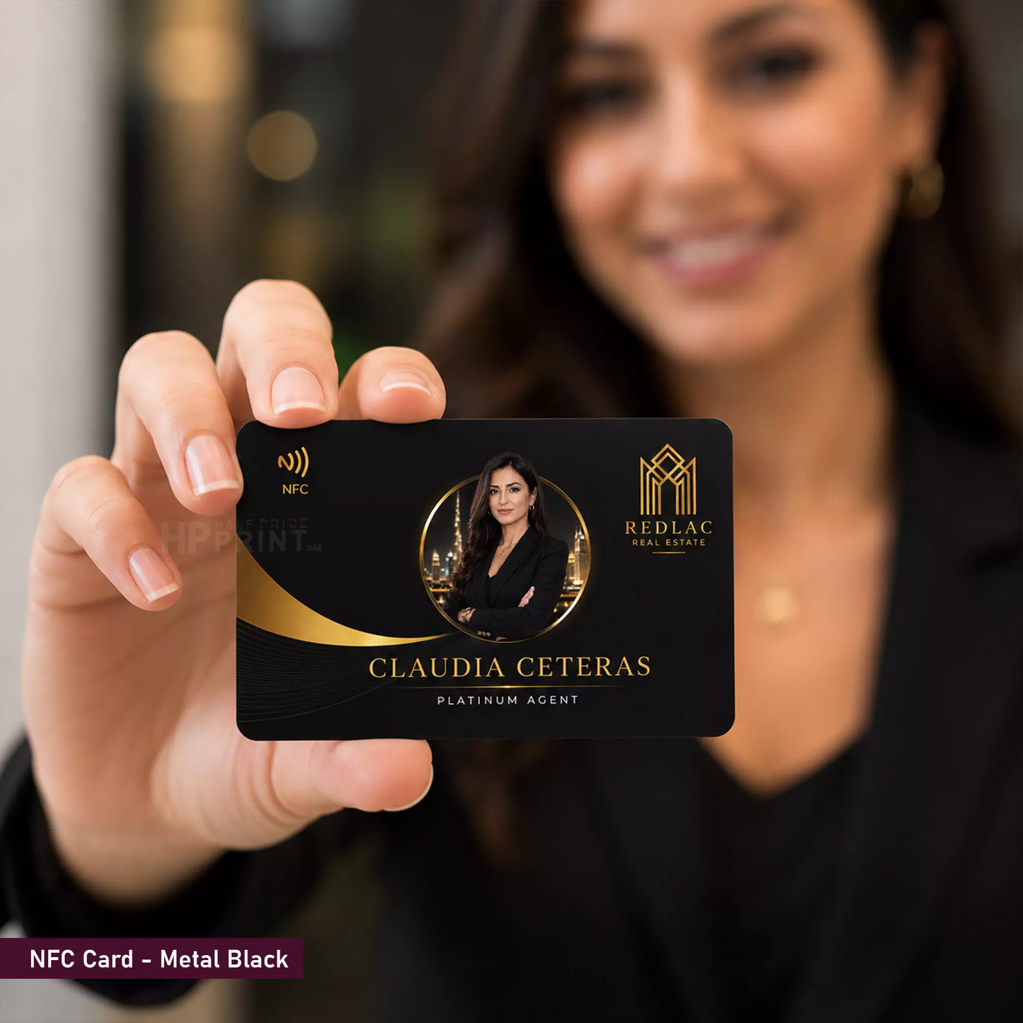 A person in a black outfit holds up a custom metal black NFC card featuring a circular photo of themselves, gold accents, and the text 'CLAUDIA CETERAS' and 'REDLAC REAL ESTATE'. The background is heavily blurred, showing the person's face and upper body.