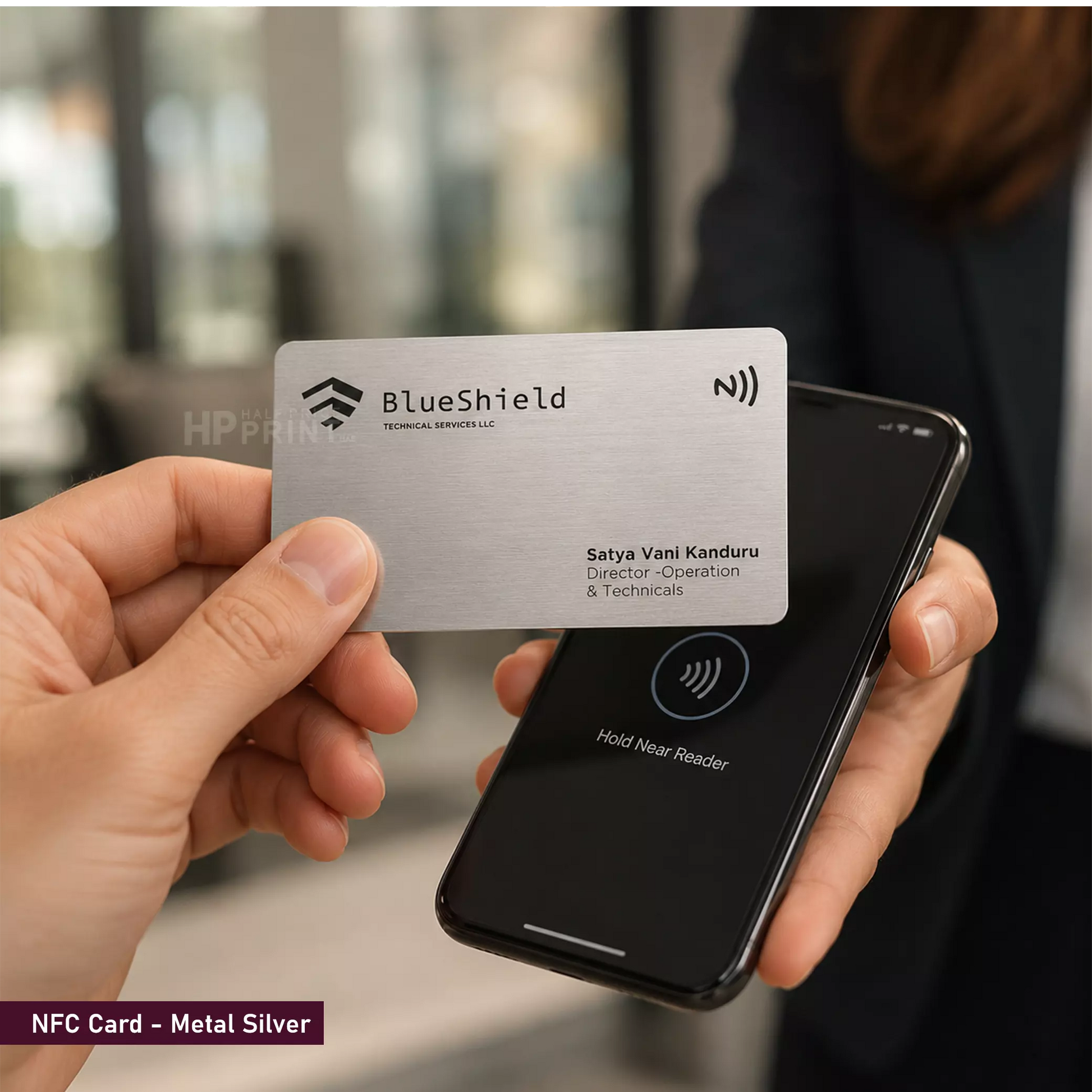 A hand holds a metallic silver NFC card branded BlueShield in front of a smartphone showing a wireless payment interface. The background is a softly blurred indoor setting with a person present, making this a lifestyle shot.