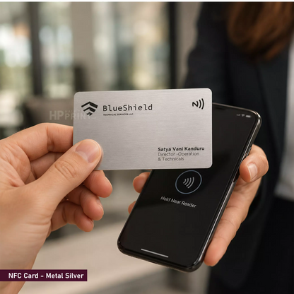 A hand holds a metallic silver NFC card branded BlueShield in front of a smartphone showing a wireless payment interface. The background is a softly blurred indoor setting with a person present, making this a lifestyle shot.