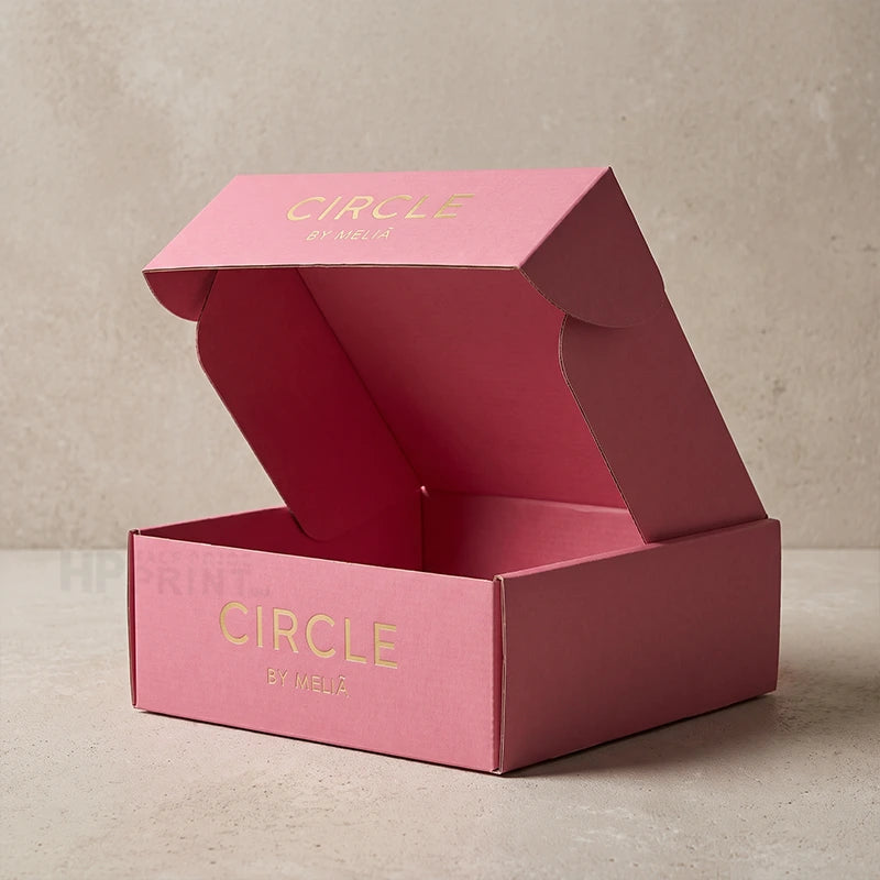 An open pink cardboard gift box with gold foil lettering that reads 'CIRCLE by Mabel' on the inside lid and front panel, set against a neutral textured beige background.