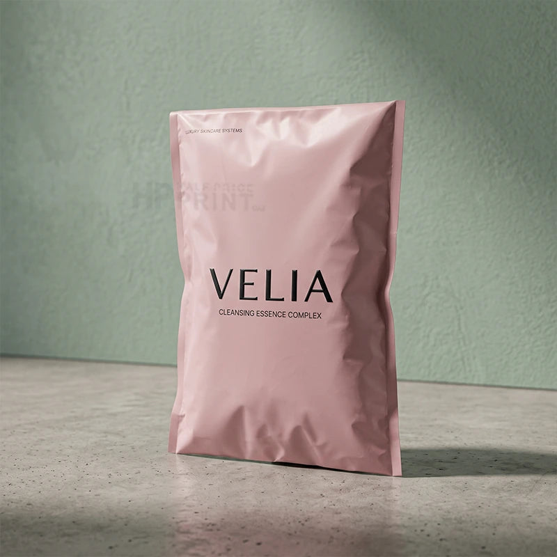 A pink foil compostable of VELIA Cleansing Essence Complex stands upright on a gray surface against a textured sage green wall.