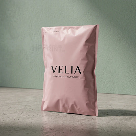 A pink foil compostable of VELIA Cleansing Essence Complex stands upright on a gray surface against a textured sage green wall.