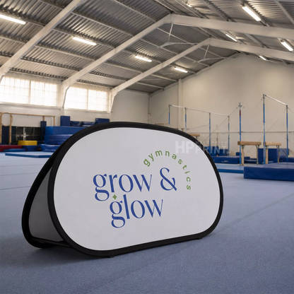 A pop-up foldable A-frame banner with a white front panel, black border, and blue and green text reading 'grow & glow gymnastics', placed on a blue gymnastics floor mat inside a large indoor gymnasium.