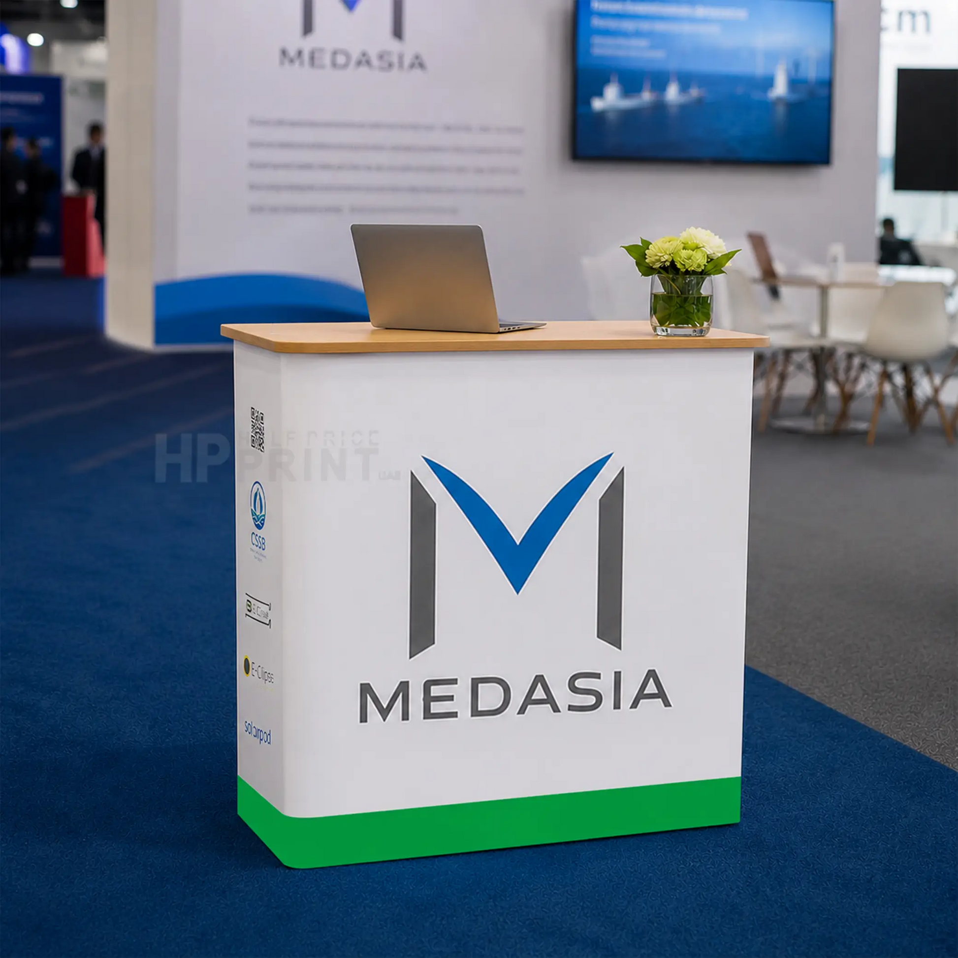 A white Medasia branded reception Fabric Pop Up Counter with a light wood top, green base, and large blue-and-gray logo, shown in a conference or exhibition setting with a laptop on top and a blurred booth background.