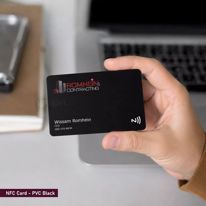 A hand holds a black PVC NFC card with the Romhein Contracting logo and contact details, with a blurred laptop in the background.