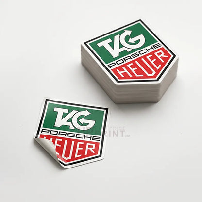 A stack of hexagonal pvc stickers and one individual sticker showing the TAG Heuer logo, displayed on a clean white background.