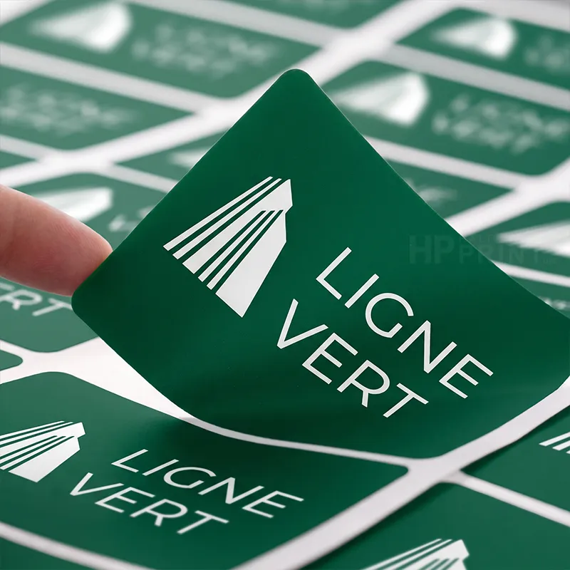 A close-up of a green square sticker being peeled from a sheet, showing the white text 'LIGNE VERT' and a white leaf-like logo. A finger is visible on the left, and more identical stickers are arranged in the background.