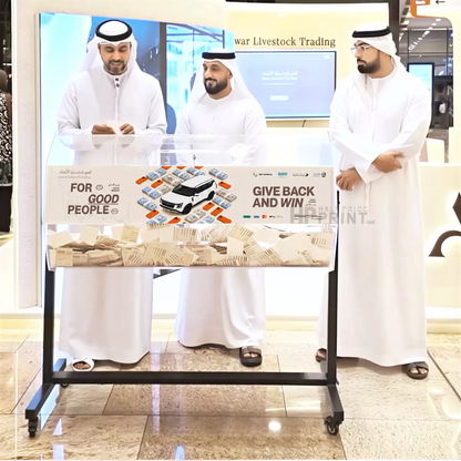 Three men in white traditional attire standing behind a counter with promotional materials in a mall setting.