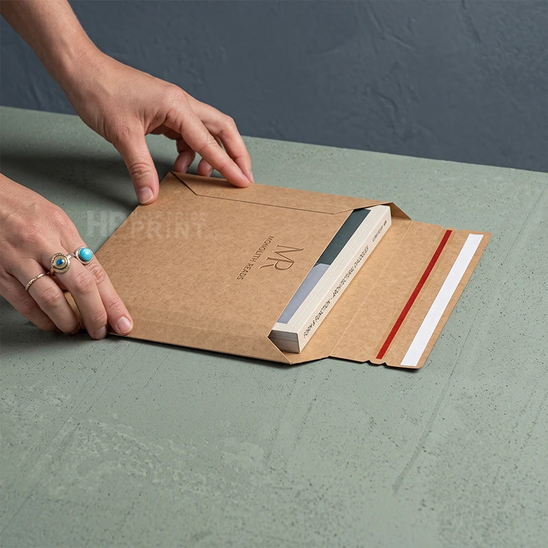 A person's hands are shown opening a brown rigid mailer envelope on a textured sage green tabletop. The envelope has a printed logo and a white strip with a red stripe on the flap.
