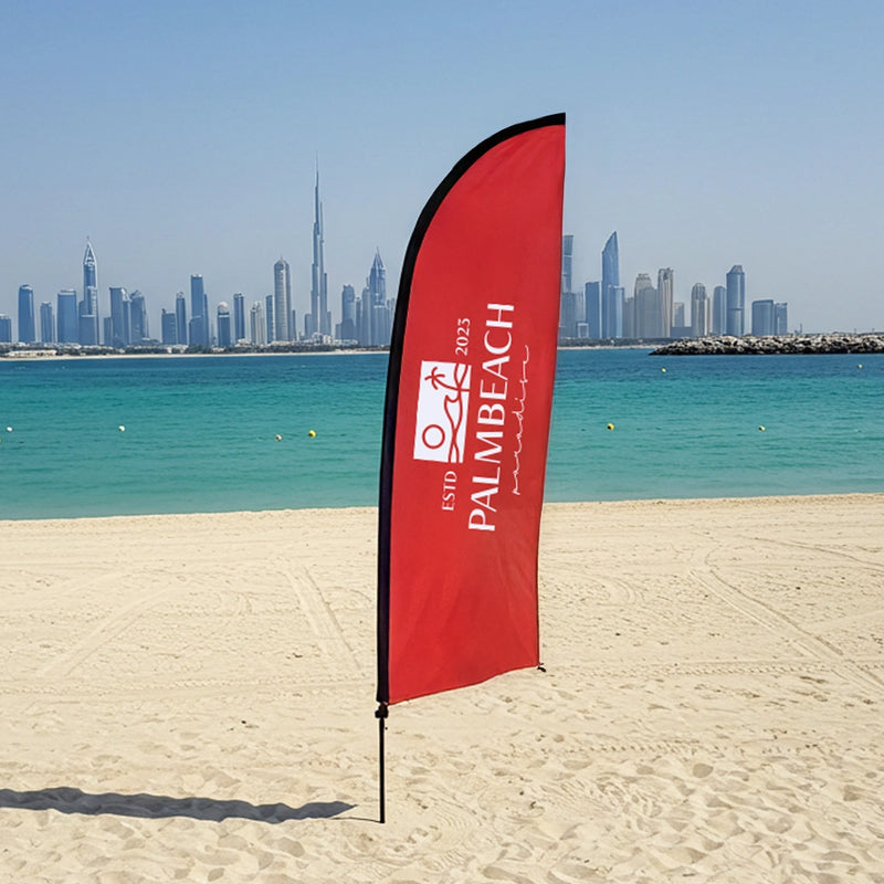 Red beach flag with 'Palm Beach' branding on a sandy beach with a city skyline in the background.