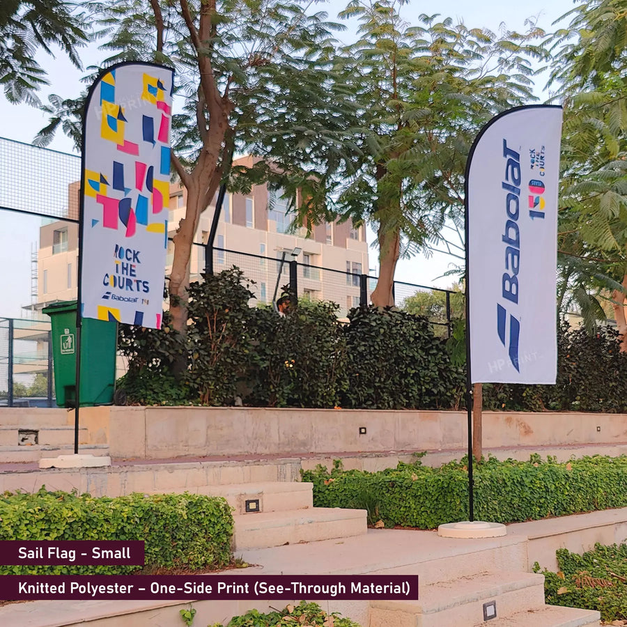 Two sail flags, one with colorful design and the other with 'Babolat' branding, in an outdoor setting.