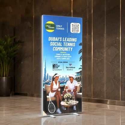 A snapfold backlist standee displaying a blue promotional graphic for the Dubai's Leading Social Tennis Community, set in a modern lobby with marble walls and a potted plant.