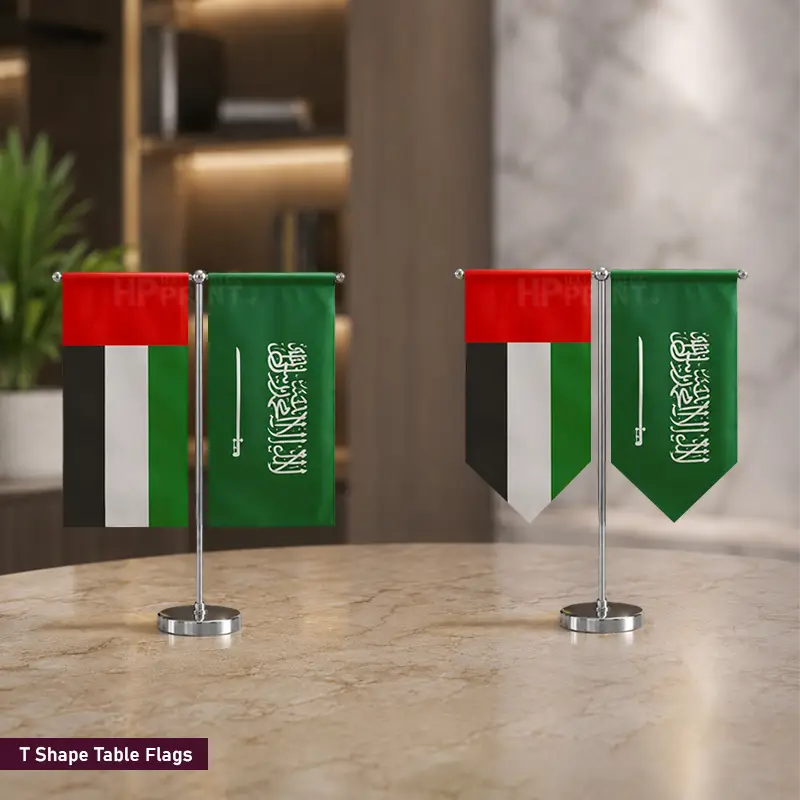 Two small table flags on silver stands displayed on a marble tabletop in an indoor setting. Each flag shows the Saudi Arabian flag design with a red, black, white, and green vertical layout, plus a pennant shape on one flag.