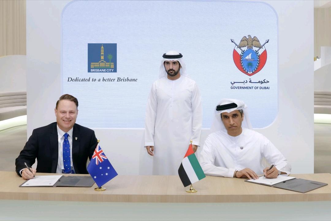 Three men at a table with flags of Australia and Dubai, behind them are two organizations' logos.