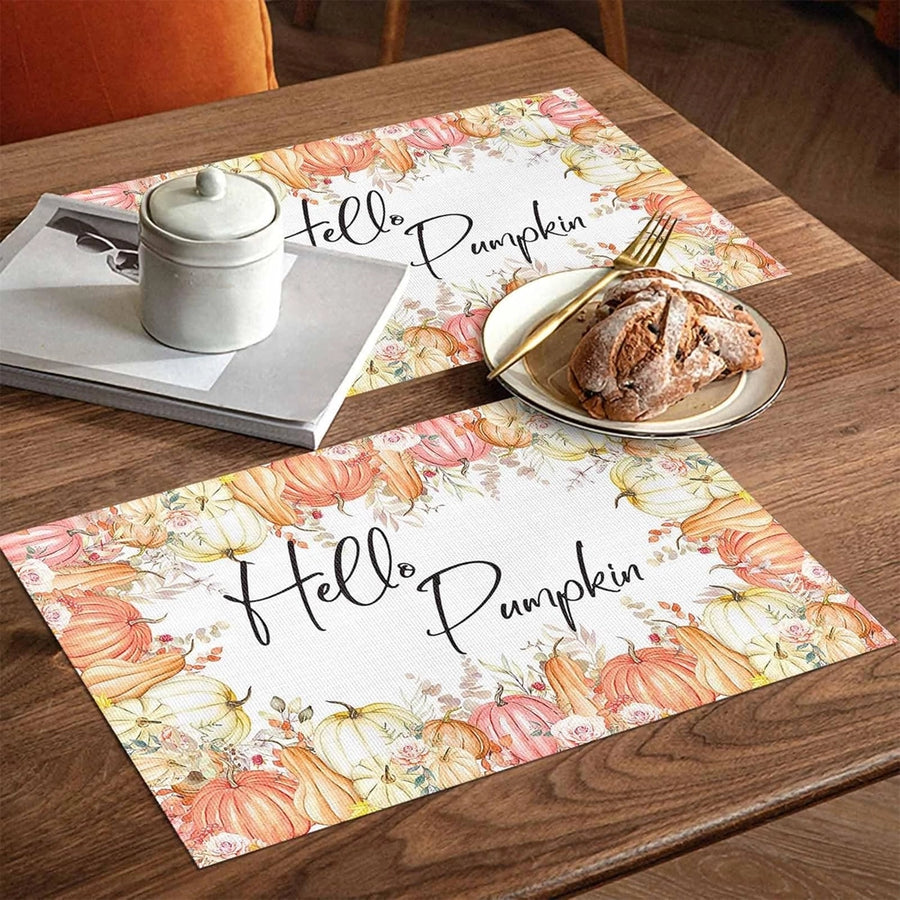 Autumn-themed placemats on a wooden table featuring pumpkins and fall leaves with the text “Hello Pumpkin,” set with a dessert plate, fork, candle jar, and napkins.