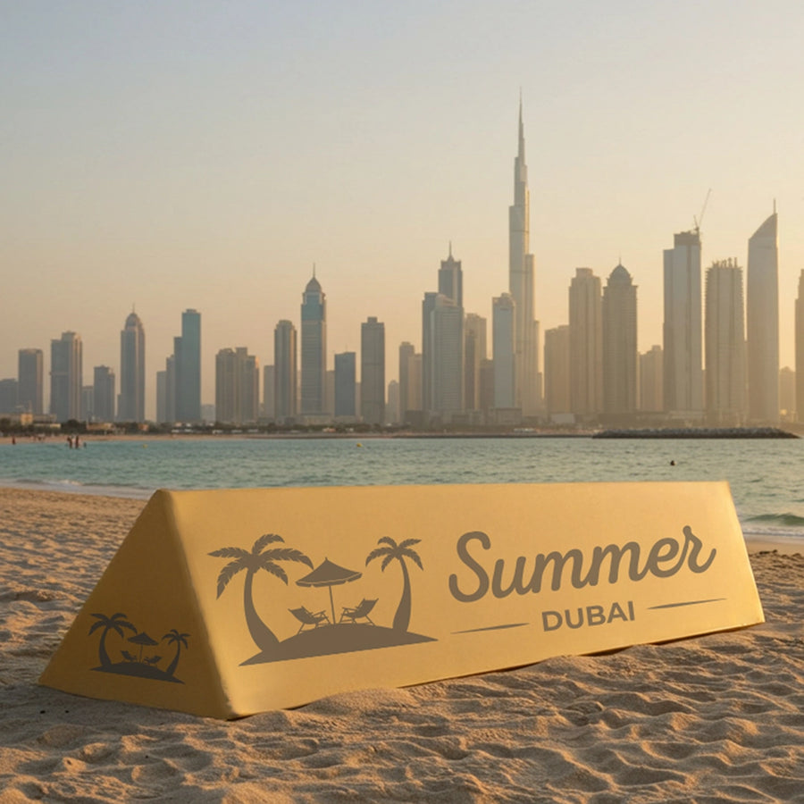 A triangular yellow A-frame advertising board with the text 'Summer Dubai' printed on it, set up on a beach with a city skyline in the background.