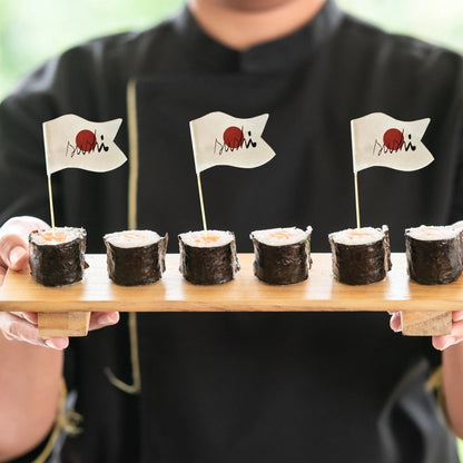 A person holding a wooden board with sushi rolls topped with tooth pick flags featuring a white and red design, presumably a logo.