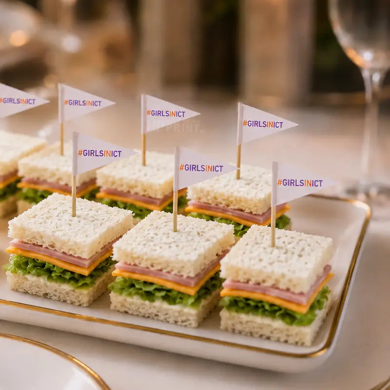 A white rectangular serving platter with a gold rim holds several small open-faced sandwiches on white bread, layered with lettuce, ham, and cheese. Each sandwich is topped with a small white toothpick flags labeled #GIRLSINICT, set in a softly blurred dining or event environment.