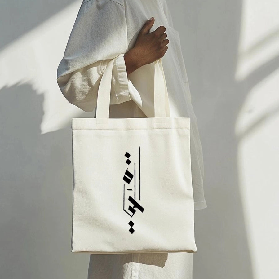 Cream canvas tote bag worn over the shoulder, featuring a vertical black Arabic calligraphy design on the front, photographed against a soft neutral background.