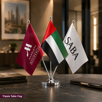 A small tabletop flag set on a dark marble desk, featuring a Marriott Residence flag, a UAE flag, and a white flag with the text SABA. The flags are mounted on a silver metal stand.