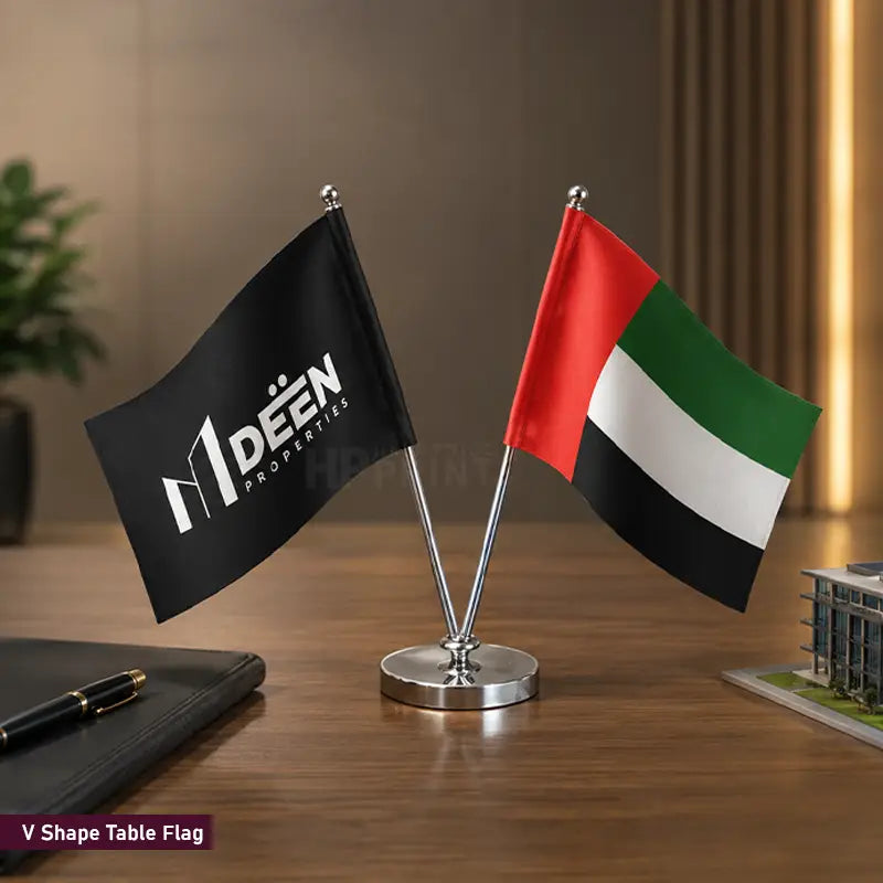 A V-shaped table flag set on a wooden desk, featuring a black flag with the 'V Deen Properties' logo on one side and the United Arab Emirates flag on the other, mounted on a silver metal stand.