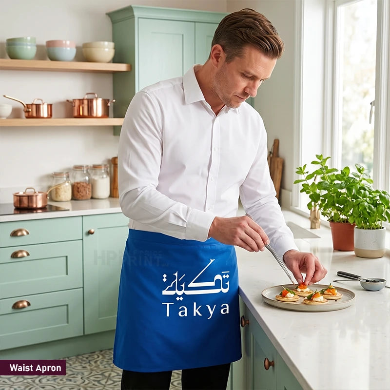 A man in a white shirt stands in a kitchen wearing a bright blue waist apron with white Arabic calligraphy and the brand name 'Takya'. He is preparing food on a plate on the kitchen counter.