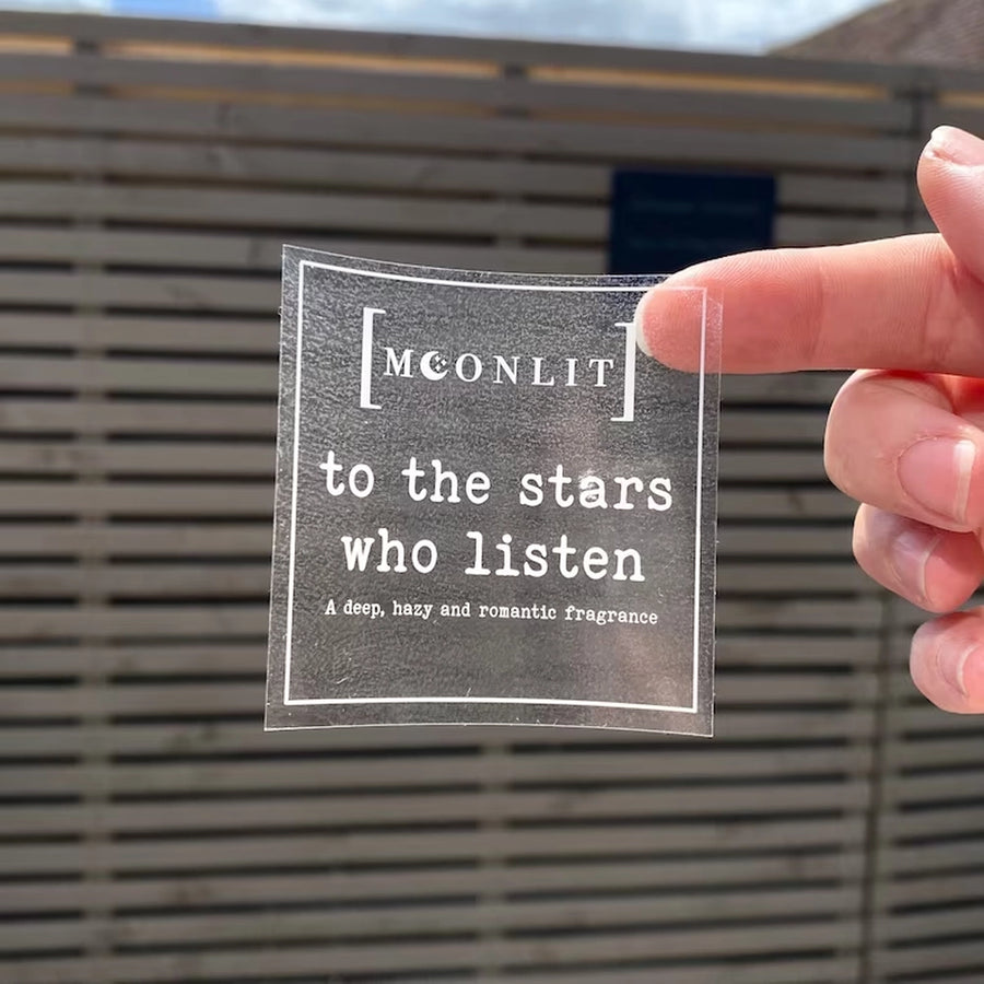 A clear square sticker with white text that reads: 'MOCNLI to the stars who listen - a deep, sad and romantic fragrance'.