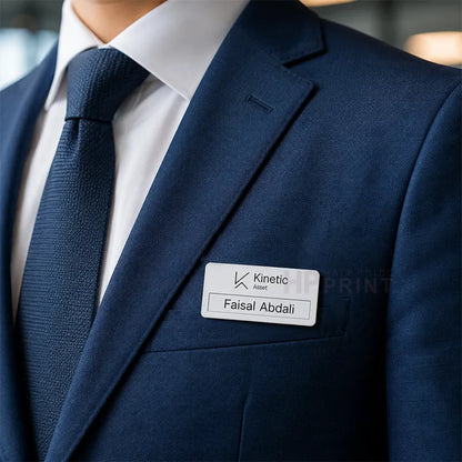 A close-up of a navy blue suit jacket with a white dress shirt and matching navy tie, featuring a white rectangular name tag on the chest pocket that reads 'Kinetic Asset' and 'Faisal Abdali'.